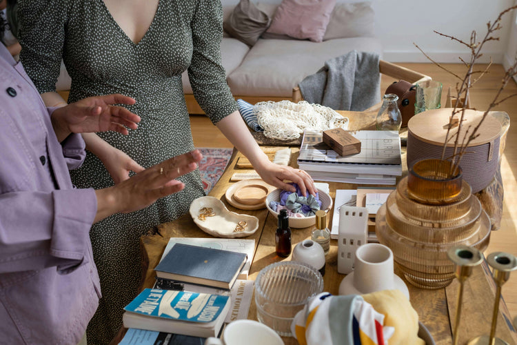 Women organizing decorative items on a table with wicker baskets, books, and pottery in a stylish living room setting.