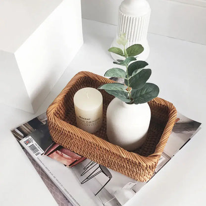 Panier de rangement en osier tressé avec une bougie blanche et un vase de feuilles vertes sur une table blanche.