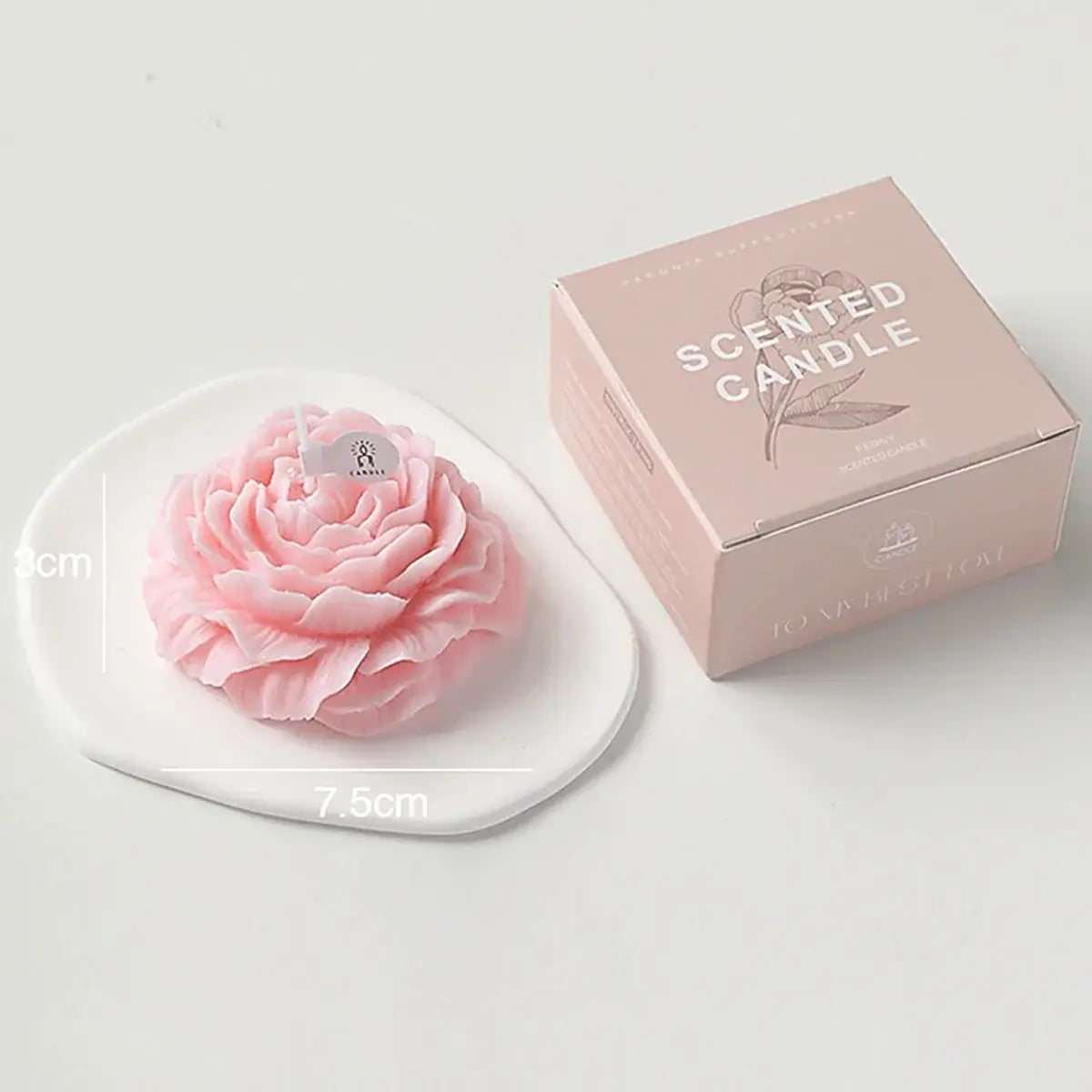 Bougie parfumée en forme de rose rose sur une assiette blanche avec boîte d'emballage rose, dimensions indiquées 7.5 cm x 3 cm.