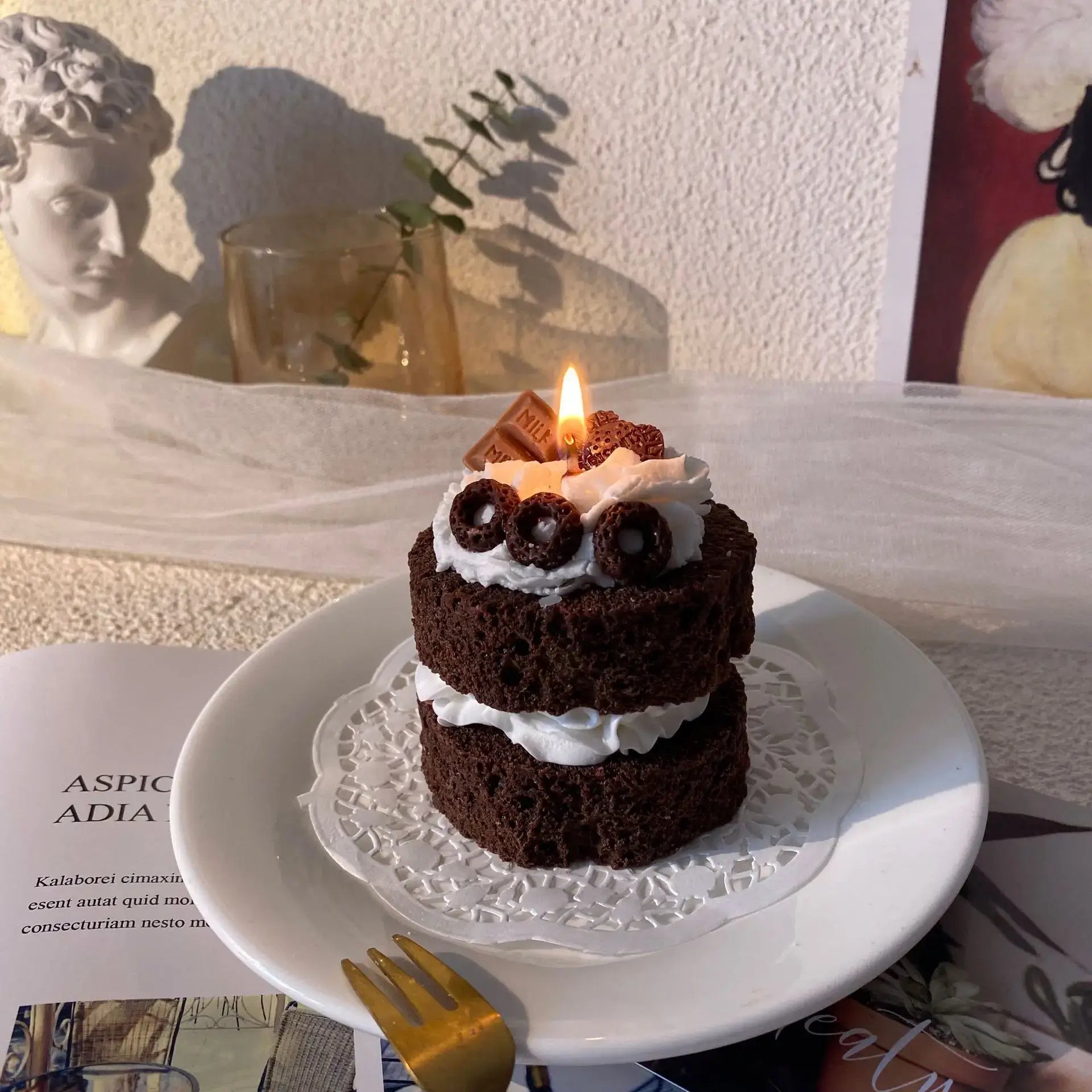 Handmade chocolate cake-shaped scented candle with whipped cream design on a white plate in a cozy, stylish home setting.