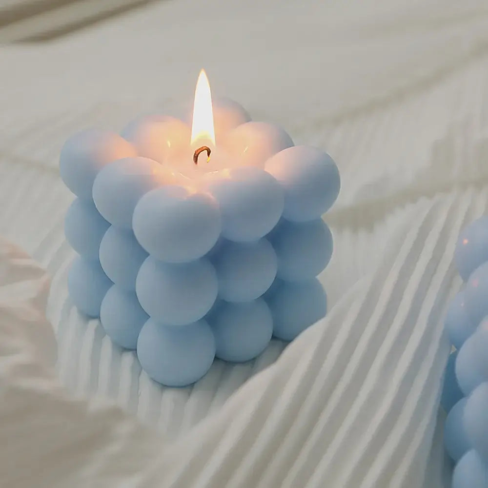 Soy wax Rubik's cube-shaped scented candle with blue bubbles burning on white fabric.