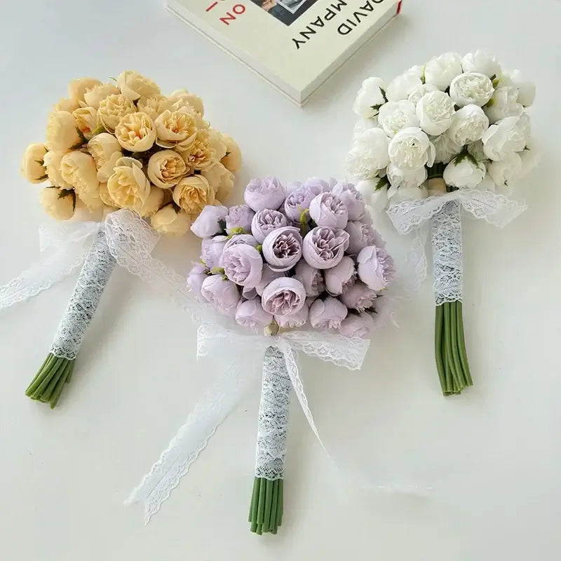 Three silk peony bouquets in yellow, lavender, and white colors, tied with lace ribbons, displayed on a table with a book in the background.