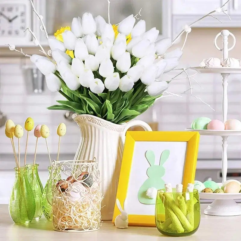 Elegant white silk tulip bouquet in a ceramic vase on a kitchen table, surrounded by Easter decorations including pastel-colored eggs and bunny-themed items.