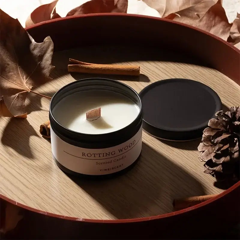 Bougie parfumée en pot noir mat avec couvercle ouvert, fabriquée à partir de cire de soja, posée sur une surface en bois entourée de feuilles sèches et de bâtons de cannelle.