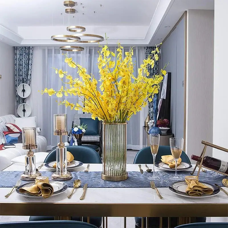 Elegant yellow orchid bouquet in tall vase on dining table with blue table runner and gold-accented dinnerware in stylish dining room.