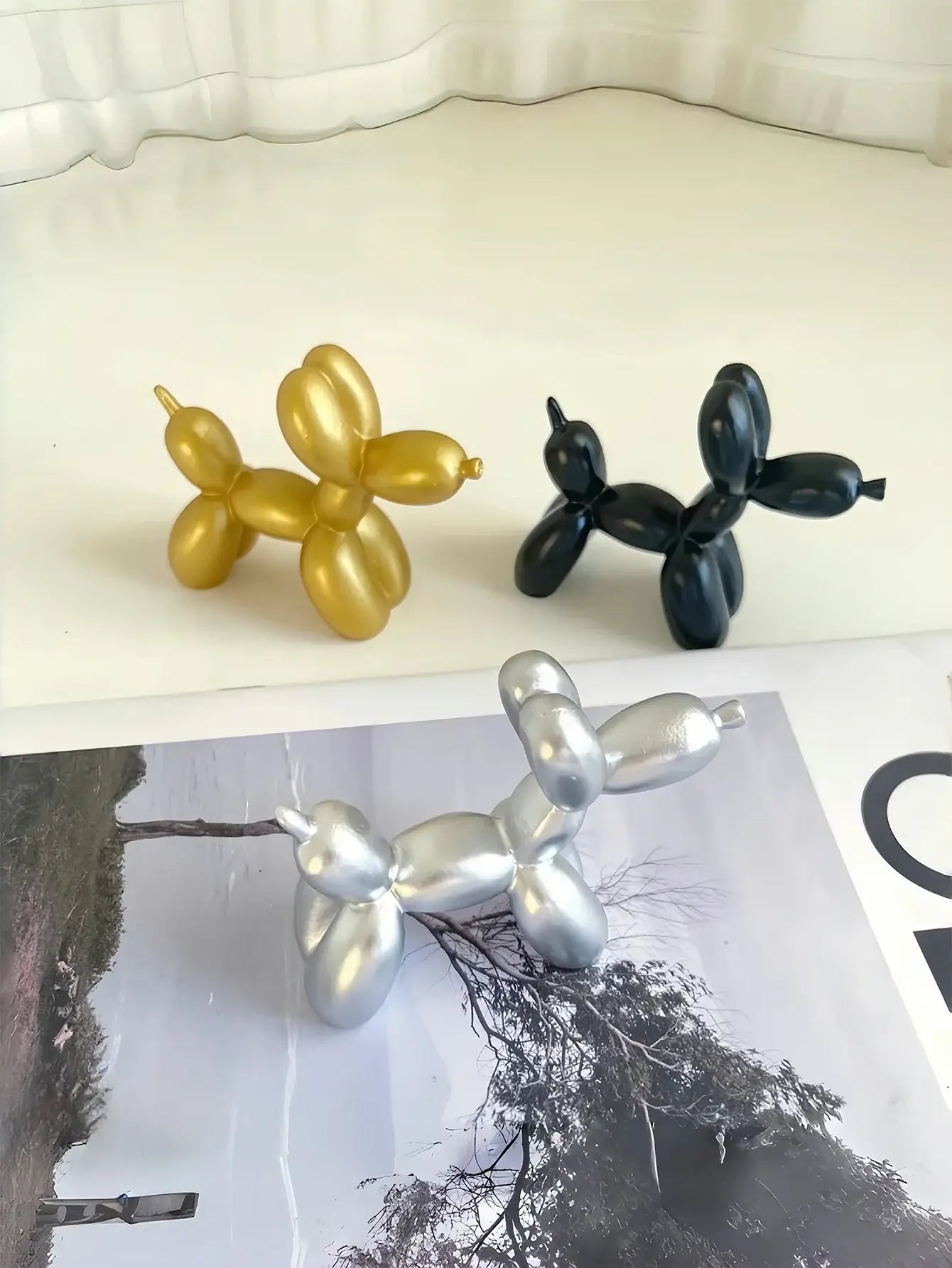 Trois statuettes de chien en ballon en résine, dorée, noire et argentée, sur une table avec un magazine, décorations ludiques pour intérieur.