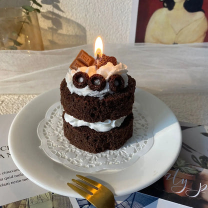 Bougie parfumée gâteau en forme de cake au chocolat avec décoration gourmande, allumée sur petite assiette blanche.