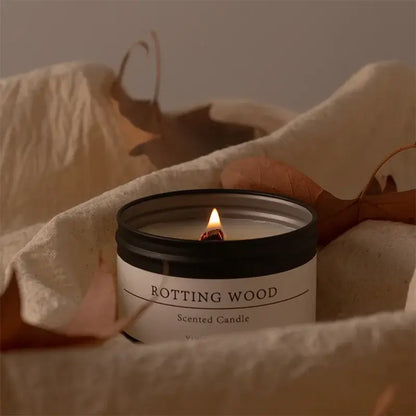 Bougie parfumée en pot noir mat, senteur bois pourri, en cire de soja allumée, entourée de feuilles sur une couverture beige