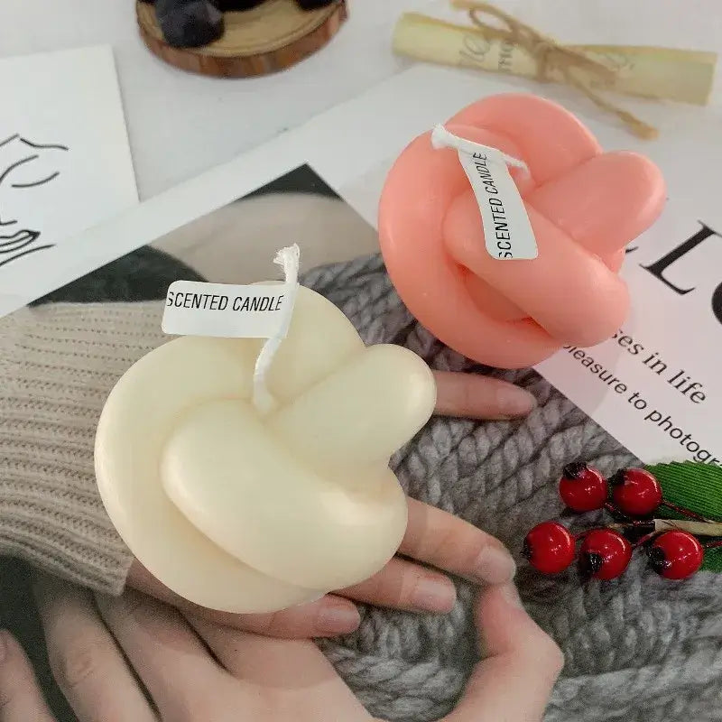 Bougies parfumées géométriques en forme de nœud, blanc et rose, sur une table avec des décorations et un magazine.