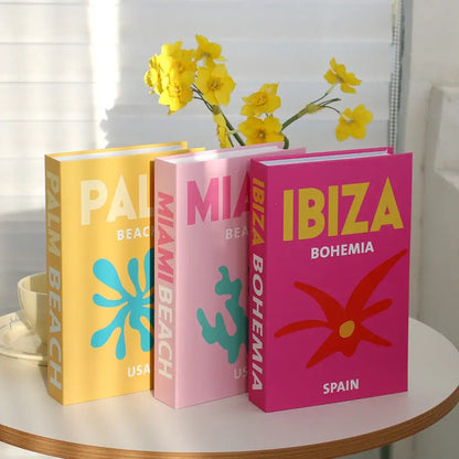 Decorative faux books with colorful covers labeled "PALM BEACH," "MIAMI BEACH," and "IBIZA BOHEMIA" on a table with yellow flowers.