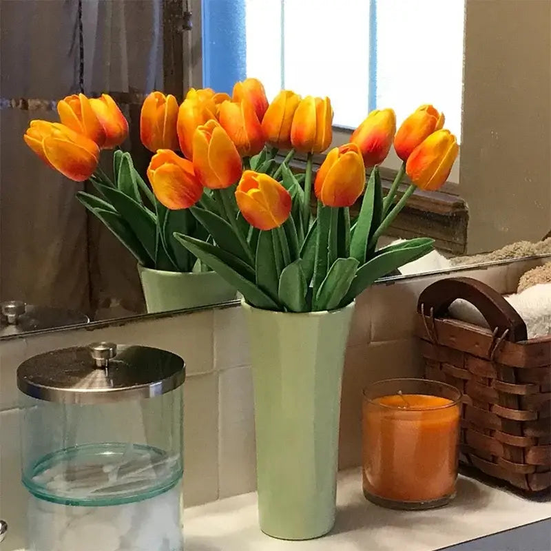 Artificial orange tulip bouquet in a green vase on a bathroom counter with a candle and a wicker basket.