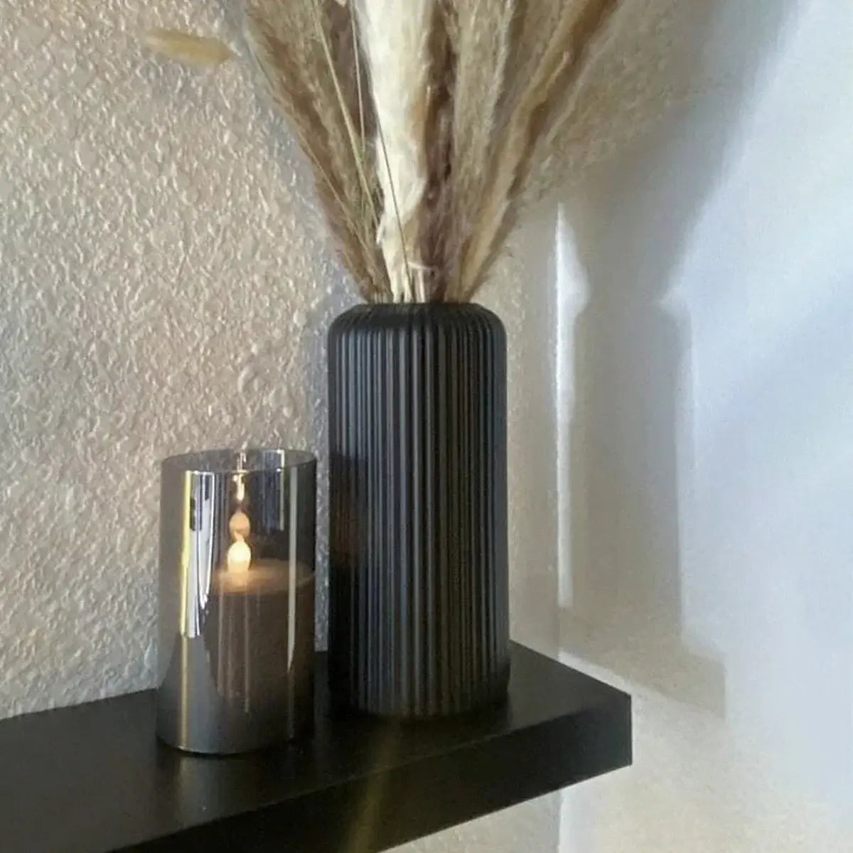 Dark striated ceramic-effect vase with dried flowers beside a silver candle holder on a black shelf