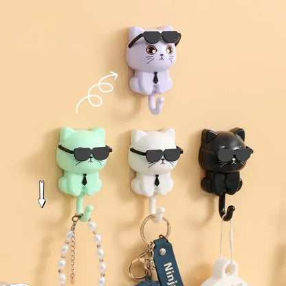 Quatre crochets porte-accessoires en forme de chat avec lunettes de soleil en noir, blanc, violet, et vert accrochés à un mur beige.