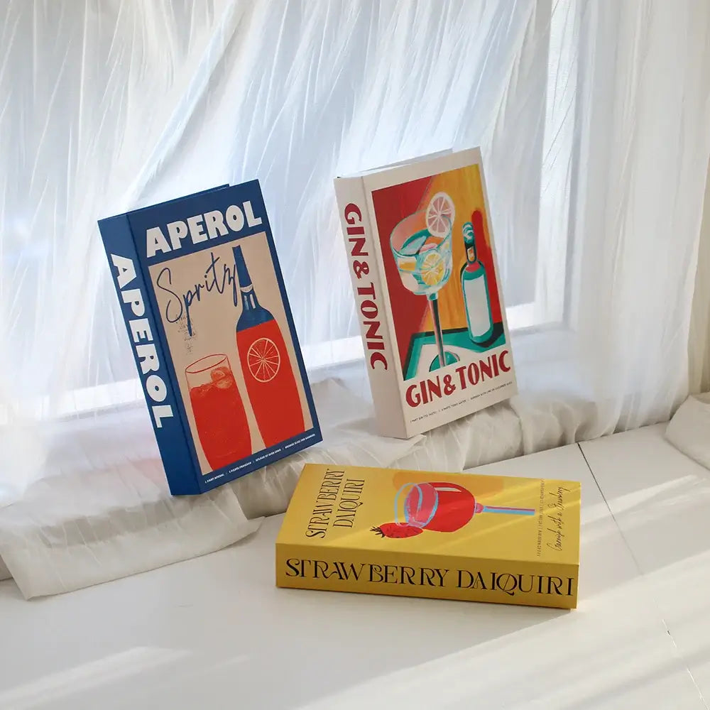 Decorative faux books with colorful cocktail-themed covers on a white windowsill, featuring Aperol, Gin & Tonic, and Strawberry Daiquiri.