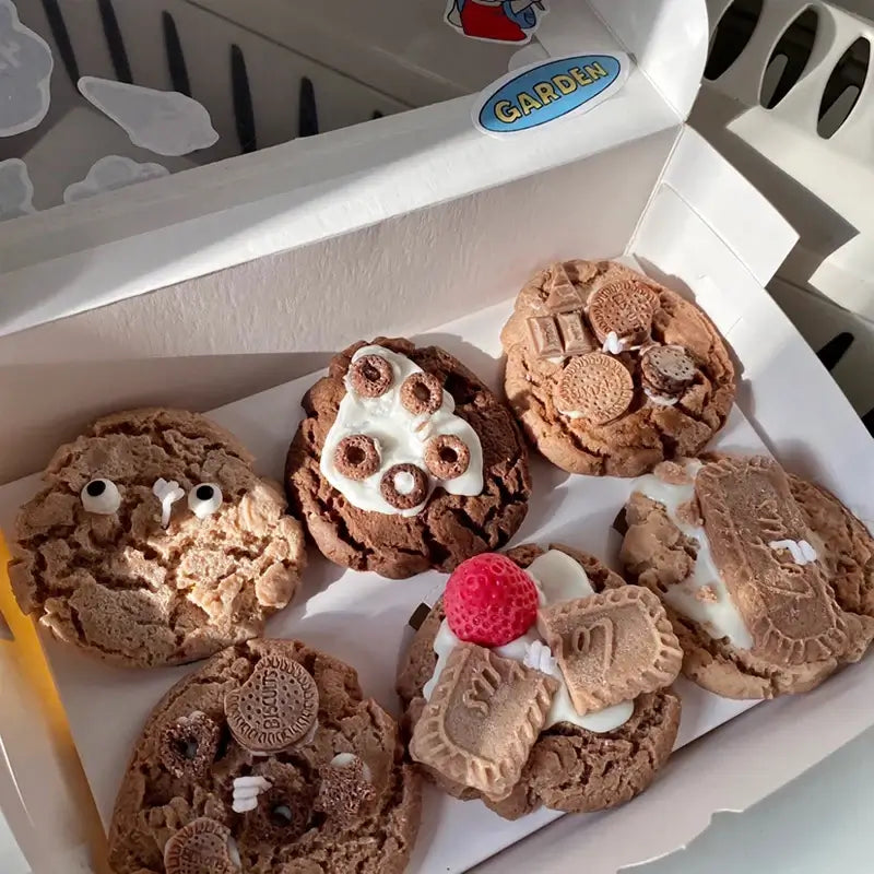 Assortment of uniquely decorated cookies resembling various designs in a white box, displayed in sunlight.