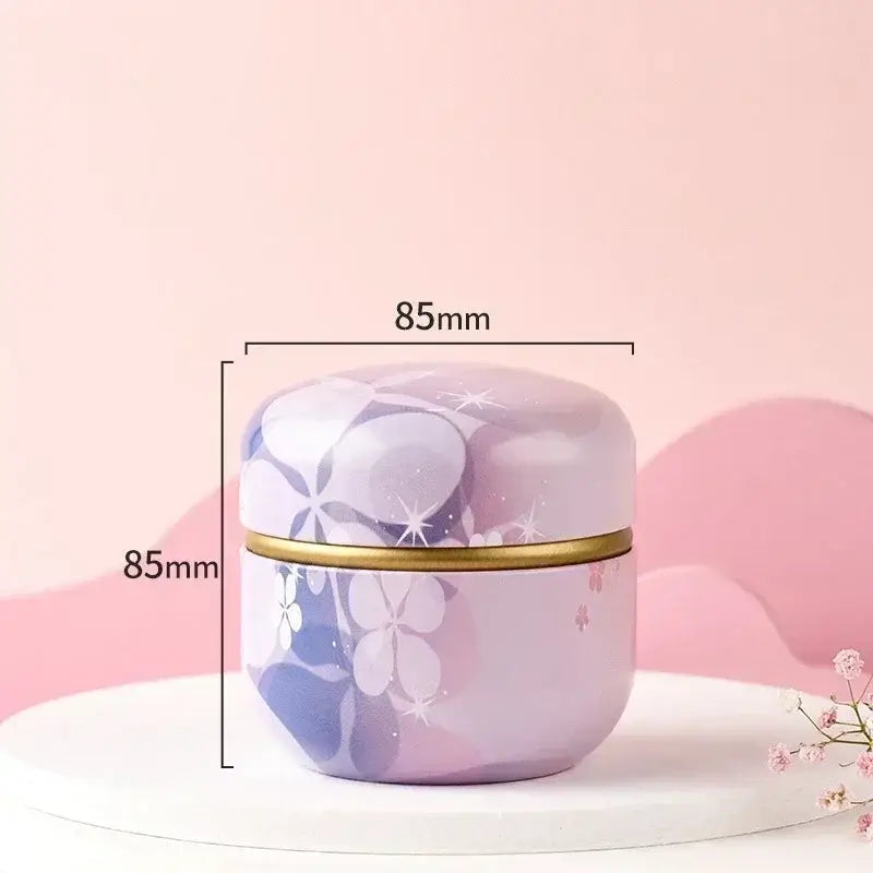 Boîte de rangement en métal décoré avec des fleurs violettes et blanches, 85mm de largeur et hauteur, sur une table blanche avec fond rose.