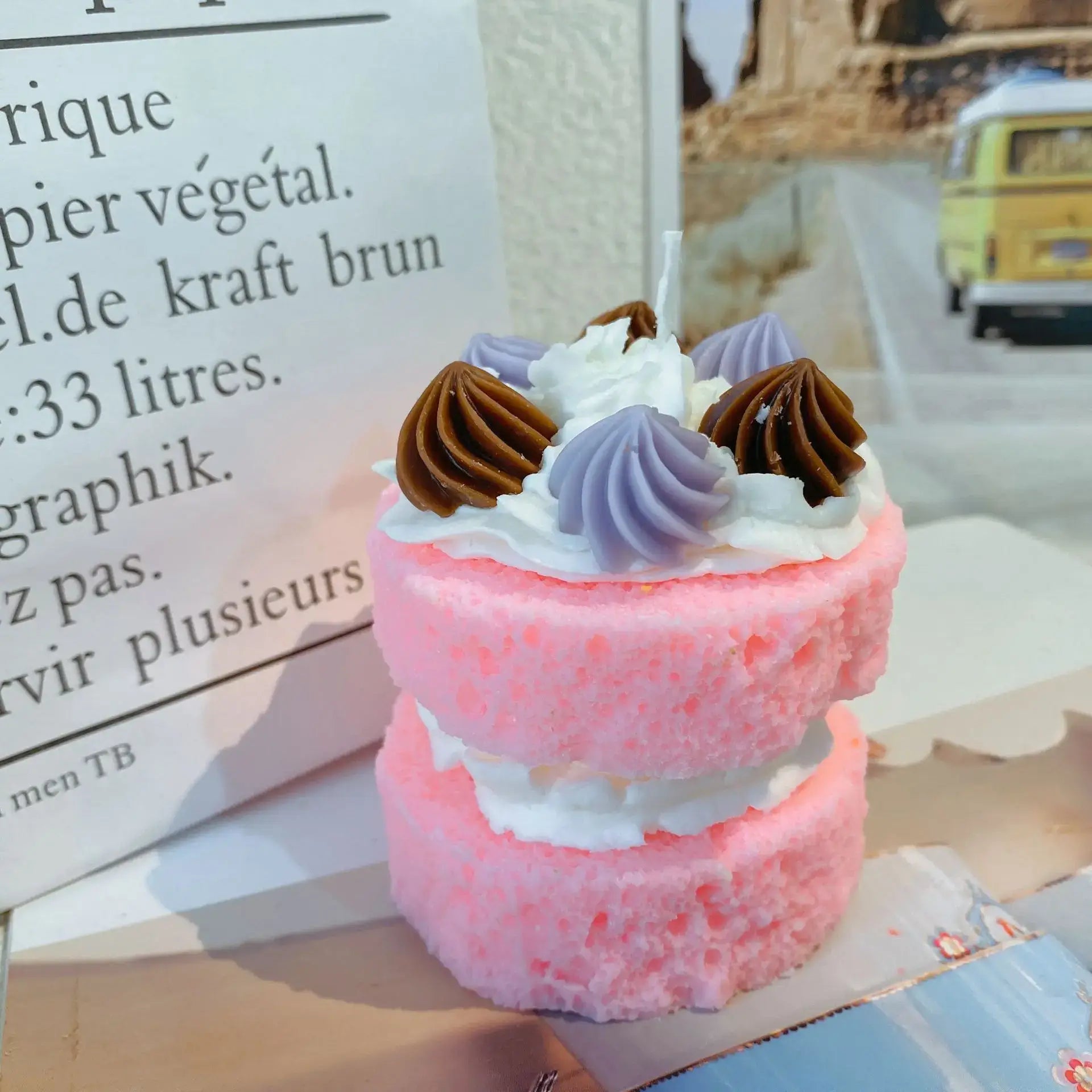 Bougie parfumée gâteau rose avec déco de crème et glaçage violet et marron, sur fond d'affiche et paysage en intérieur.