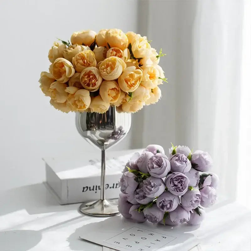 Bouquet de fleurs de pivoines en soie réalistes, une jaune dans un vase argenté et une violette posée sur une table blanche.